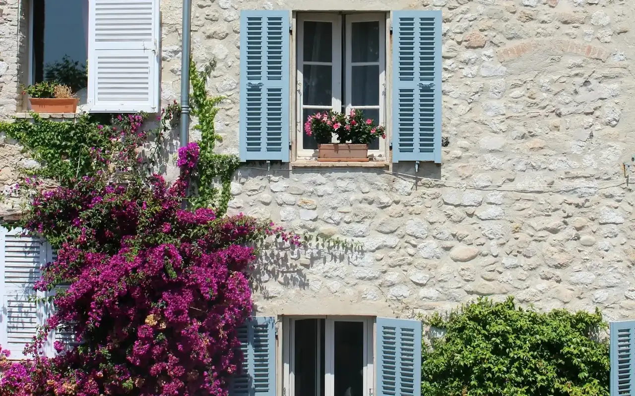 Façade en pierre aux volets bleus, fleurie de bougainvilliers